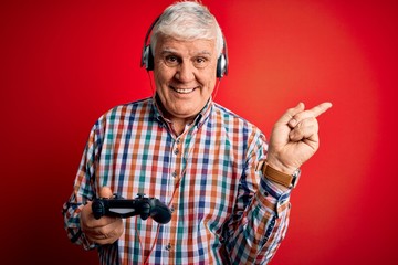 Senior handsome hoary gamer man playing video game using joystick and headphones very happy pointing with hand and finger to the side