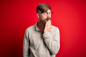 Handsome Irish redhead man with beard wearing casual sweater and glasses over red background bored yawning tired covering mouth with hand. Restless and sleepiness.