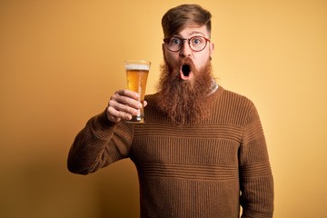 Irish redhead man with beard drinking a glass of refreshing beer over yellow background scared in...