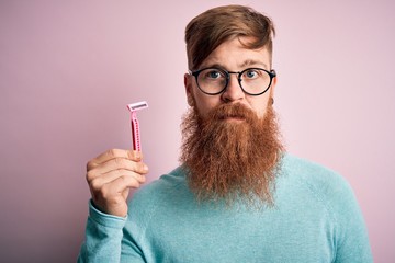 Redhead Irish man with beard holding beauty razor for shaving and skin care over pink background...