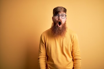 Handsome Irish redhead man with beard wearing glasses over yellow isolated background afraid and shocked with surprise and amazed expression, fear and excited face.