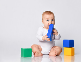 Baby boy in bodysuit, barefoot. He is playing with multi-colored toys, sitting on floor isolated on white. Close up, copy space