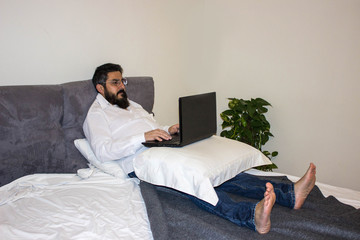 handsome bearded man wearing formal white shirt and jeans on the bed with laptop in modern room. Remote work concept. Man surfing internet or working online.
