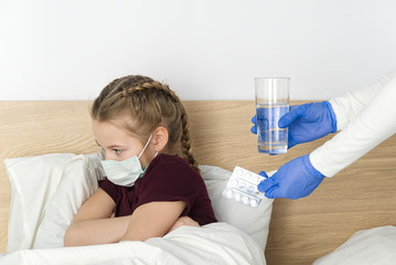 A young girl lying on the bed is given pills and a glass of water. masked girl for protection against viral diseases. concept of quarantine and treatment for viral diseases, colds and coronavirus