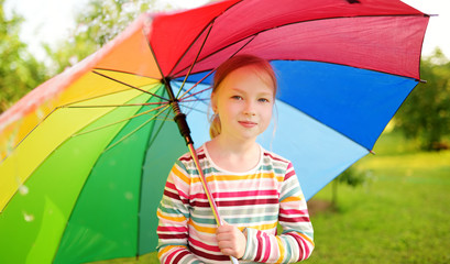 Cute girl holding colorful rainbow umbrella on rainy summer day. Child walking under warm rain outdoors. Outdoor summer activities for kids.
