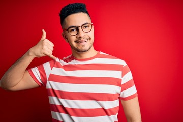 Young handsome man wearing casual striped t-shirt and glasses over isolated red background smiling doing phone gesture with hand and fingers like talking on the telephone. Communicating concepts.
