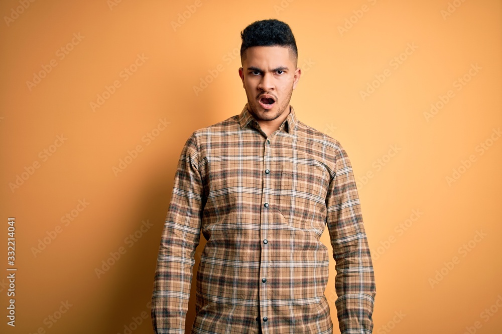 Wall mural young handsome man wearing casual shirt standing over isolated yellow background in shock face, look