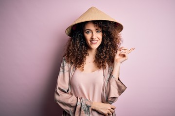 Young beautiful woman with curly hair and piercing wearing traditional asian conical hat with a big smile on face, pointing with hand and finger to the side looking at the camera.