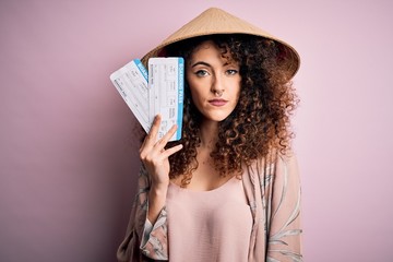 Young beautiful woman with curly hair and piercing wearing asian hat holding boarding pass with a confident expression on smart face thinking serious