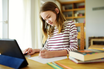 Smart preteen schoolgirl doing her homework with digital tablet at home. Education and distance...