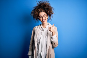 Fototapeta na wymiar Young beautiful businesswoman with curly hair and piercing wearing jacket and glasses doing happy thumbs up gesture with hand. Approving expression looking at the camera showing success.