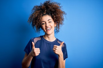 Fototapeta na wymiar Young beautiful woman with curly hair and piercing wearing casual blue t-shirt success sign doing positive gesture with hand, thumbs up smiling and happy. Cheerful expression and winner gesture.
