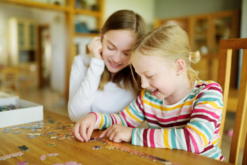 Fototapeta premium Cute young girls playing puzzles at home. Children connecting jigsaw puzzle pieces in a living room table. Kids assembling a jigsaw puzzle. Fun family leisure.
