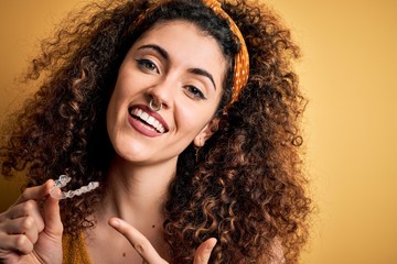 Young beautiful woman with curly hair and piercing holding dental aligner orthodontic very happy...