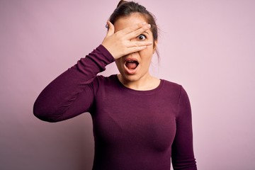 Beautiful young woman wearing casual bun hairstyle over pink isolated background peeking in shock covering face and eyes with hand, looking through fingers with embarrassed expression.