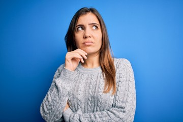 Beautiful young woman wearing casual wool sweater standing over blue isolated background with hand on chin thinking about question, pensive expression. Smiling with thoughtful face. Doubt concept.