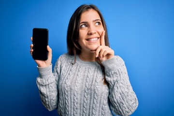 Young woman showing blank smartphone screen over blue isolated background serious face thinking...