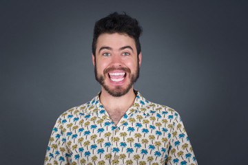 Emotive young blond man wears casual clothes, happy and excited to celebrate anniversary. Happy young man has good mood stands against gray wall.