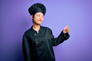 Young beautiful chinese chef woman wearing cooker uniform and hat over purple background Looking proud, smiling doing thumbs up gesture to the side