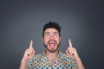 Charming carefree man with positive expression, points up with both index fingers, dressed in...
