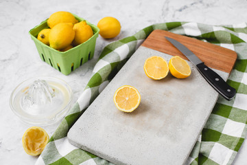 Fresh Lemons in a Green Produce Basket on a White Marble Countertop; Some Cut Open on a Cement Cutting Board; Green and White Checked Kitchen Towel in Background; Lemon Juicer with Juice Beside