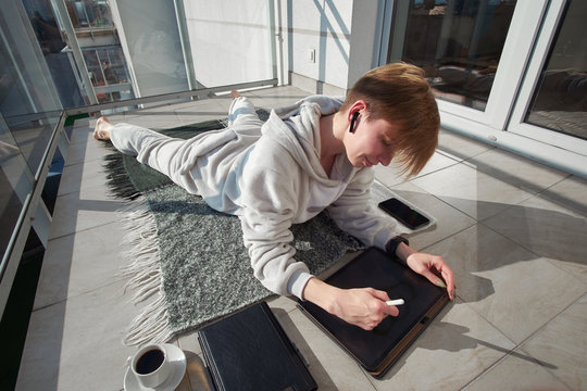 Shorthair redhead girl digital art illustrator weared in pijamas lies on sunny balcony, listening to music in earphones, drinking coffee and working on tablet
