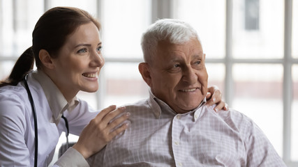 Fototapeta premium Young female nurse wearing white coat hugs old man grey-haired patient smiling look at distance. Concept of eldercare, health check-up, geriatrics medicine, social worker caretaker caring about senior
