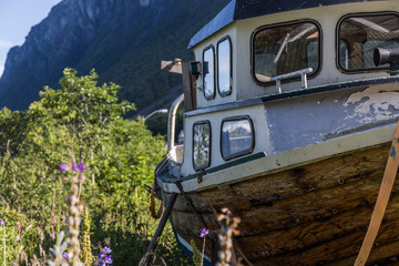 Fototapeta na wymiar old wooden boat, brown, blue and white washed in the grass, next to a deep blue Norwegian fjord, in the background we can see the mountains, clear summer day with some clouds