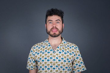 Beautiful Young caucasian man with happy and funny face smiling and showing tongue. Wearing casual clothes and standing against gray studio background.