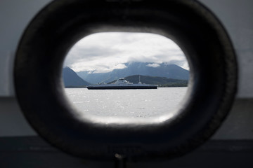 through the porthole of a ship we can see another ship sailing through a Norwegian fjord, the water is dark and the day is cloudy