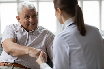 Nurse in uniform talking during day visit to old patient. Caring caregiver hold hand of 80s man...