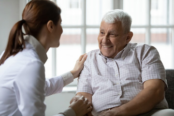 Caring nurse talks to old patient holds his hand sit in living room at homecare visit provide...