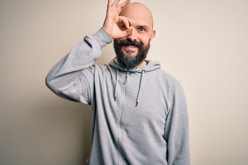 Handsome sporty bald man with beard wearing sweatshirt standing over pink background doing ok gesture with hand smiling, eye looking through fingers with happy face.