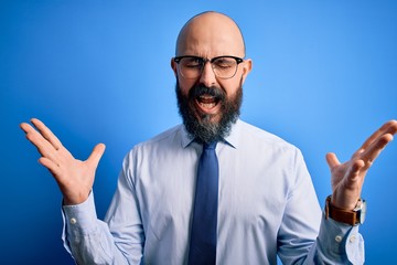 Handsome business bald man with beard wearing elegant tie and glasses over blue background celebrating mad and crazy for success with arms raised and closed eyes screaming excited. Winner concept