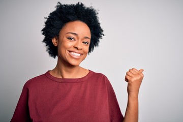 Young beautiful African American afro woman with curly hair wearing casual t-shirt standing smiling with happy face looking and pointing to the side with thumb up.
