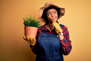 Young African American farmer woman with curly hair wearing apron holding pot with plants serious...
