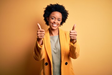 Young beautiful African American afro businesswoman with curly hair wearing yellow jacket approving doing positive gesture with hand, thumbs up smiling and happy for success. Winner gesture.