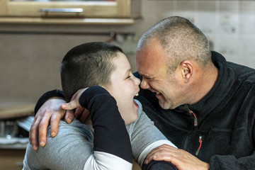 Father and son sitting at the table in the kitchen are hugging and laughing face to face.