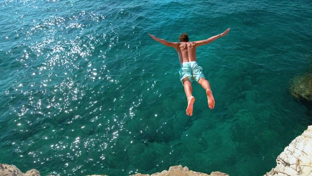 SLOW MOTION, COPY SPACE: Athletic young man jumping off a rocky ledge and into the glistening blue ocean. Male tourist on a relaxing summer vacation in Croatia does cliff diving on a sunny day.