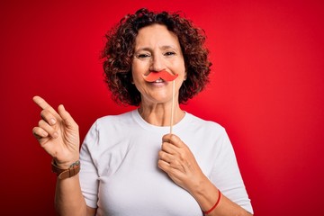 Middle age curly hair woman holding funny mustache over isolated red background very happy pointing...