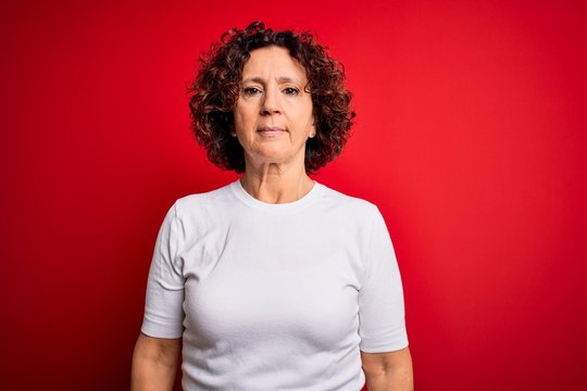 Middle age beautiful curly hair woman wearing casual t-shirt over isolated red background with serious expression on face. Simple and natural looking at the camera.