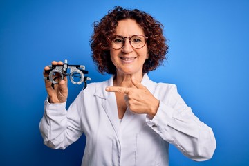 Middle age optical woman wearing coat holding optometry glasses over blue background very happy pointing with hand and finger