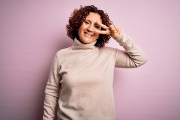 Fototapeta na wymiar Middle age beautiful curly hair woman wearing casual turtleneck sweater over pink background Doing peace symbol with fingers over face, smiling cheerful showing victory