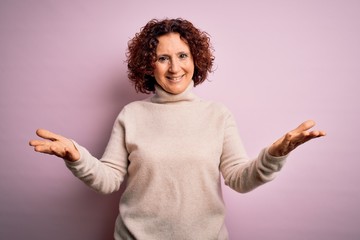 Middle age beautiful curly hair woman wearing casual turtleneck sweater over pink background smiling cheerful offering hands giving assistance and acceptance.