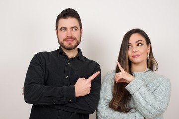 Pretty brunette couple posing on camera with tricky look and gesturing to side. Female designer presenting lovely product with index finger. Gesture concept.
