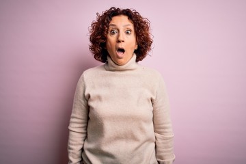 Middle age beautiful curly hair woman wearing casual turtleneck sweater over pink background afraid and shocked with surprise and amazed expression, fear and excited face.