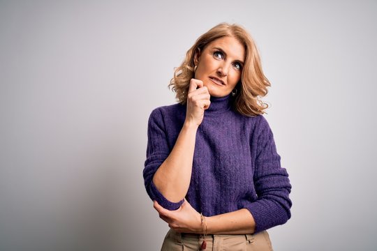 Middle age beautiful blonde woman wearing purple turtleneck sweater over white background with hand on chin thinking about question, pensive expression. Smiling with thoughtful face. Doubt concept.
