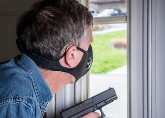 Senior man with face mask looking worried at front door and ready to defend home with a gun during...