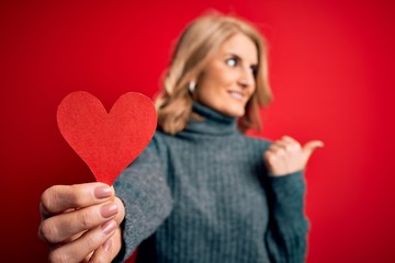Middle age beautiful blonde romantic woman holding red paper heart celebrating valentine day pointing and showing with thumb up to the side with happy face smiling