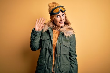 Middle age beautiful blonde skier woman wearing snow sportwear and ski goggles Waiving saying hello happy and smiling, friendly welcome gesture